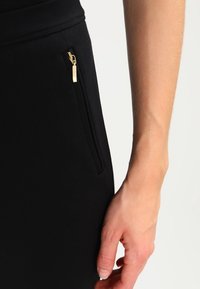 Svart tyg med en slät yta, åtsittande design, med en guld dragkedjedetalj på fickan, som visar en samtida stil.