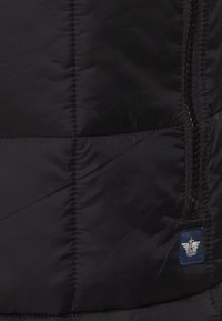 Czarna ocieplana kamizelka z pionowymi szwami, pełnym suwakiem oraz małą naszywką z logo w niebieskim kolorze z białymi detalami.