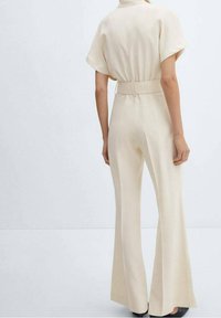 Vrouw in beige hoge taille wijde broek en een bijpassend top met korte mouwen, staand tegen een effen lichte achtergrond.