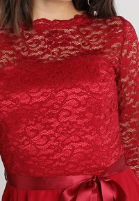 Haut en dentelle rouge à manches longues, présentant des motifs floraux intricats. Ceinture en satin avec un accent de nœud, créant une silhouette ajustée.