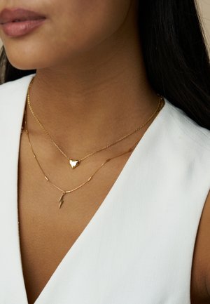 Χρυσά κολιέ στρωμένα το ένα πάνω στο άλλο: ένα μενταγιόν σε σχήμα καρδιάς και ένα μενταγιόν σε μορφή αστραπής σε λεπτές αλυσίδες, φορεμένα πάνω από ένα λευκό πουκάμισο.