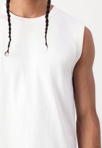 Tricou alb din bumbac fără mâneci, cu guler rotund, având o textură netedă și o cădere subtilă, completat cu două fire împletite.