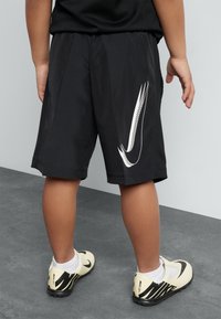 Black athletic shorts with a white Nike logo, featuring a lightweight mesh fabric and side pockets. Complemented by beige and black shoes.