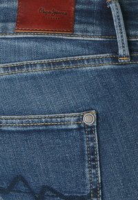 Jeans de mezclilla en azul medio con costuras doradas en contraste. Cuenta con un parche de cuero en la cinturilla y un detalle de botón de metal.