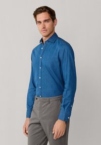 Modrá deninová košeľa s dlhým rukávom a golierom na gombíky, s úzkym strihom a kontrastnými bielymi gombíkmi, v kombinácii so šedými nohavicami.