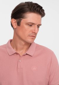 Ljust rosa polo-skorta med en texturerad tyg, två knappar vid kragen och en broderad logotyp på vänster bröst.