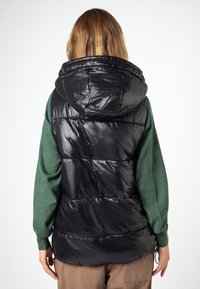 Schwarze gesteppte Weste mit Kapuze, die eine glänzende, gesteppte Textur und elastische Bündchen aufweist. Wird über einem grünen Pullover getragen, mit sichtbarer brauner Hose.