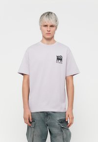 Lys lilla bomulds T-shirt, rund hals, med en sort grafik af en kat på venstre bryst. Løs pasform, korte ærmer, blød tekstur.