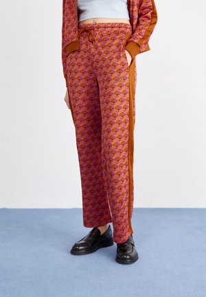 Vrouw draagt oranje en paarse wijde broek met geometrisch patroon, bruine zijstrepen en zwarte loafers, staand op een blauwe vloer.