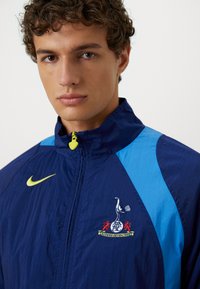 Blå og marineblå zip-up jakke med et gult Nike-logo, lyseblå detaljer og et broderet emblem med en fugl og røde løver.