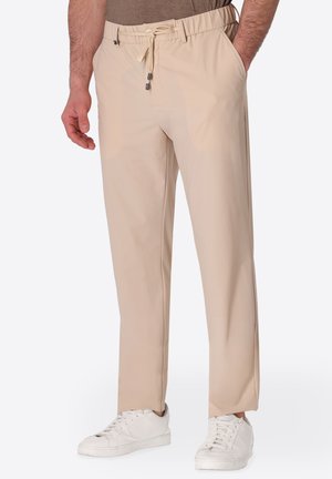 Uomo che indossa pantaloni beige con coulisse e sneakers bianche, in piedi con una mano in tasca e l'altra rilassata lungo il fianco.