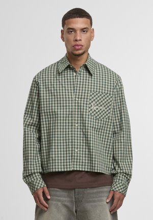 Muž oblečený v zelenej a béžovej kárované košeli s vreckom na hrudi, tmavohnedým tričkom a vyblednutými sivými džínsami, stojaci na bielom pozadí.