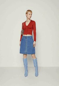 Blusa justa de manga longa vermelha com botões na frente, combinada com uma saia em A de ganga azul com fecho de botão na frente e botas azuis até ao joelho.