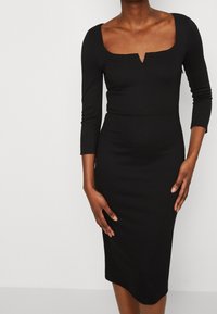 Musta bodycon-mekko, jossa on nelikulmainen pääntie ja kolmen neljänneksen hihat. Mekossa on tiukka malli, joka levenee hieman helmaa kohti.