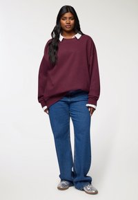 Młoda kobieta z długimi czarnymi włosami, ubrana w bordowy, oversize'owy sweter, niebieskie dżinsy, białe mankiety koszuli i srebrne trampki, stojąca na tle białym.