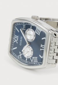 Montre-bracelet en argent avec un boîtier rectangulaire, un cadran bleu, des chiffres romains, deux sous-cadrans et un bracelet en métal maillé.