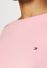 Vaaleanpunainen puuvillapaita, jossa on pyöreä pääntie, vasemmassa rinnassa pieni punaisista, valkoisista ja sinisistä langoista brodeerattu logo. Silkkinen pinta.