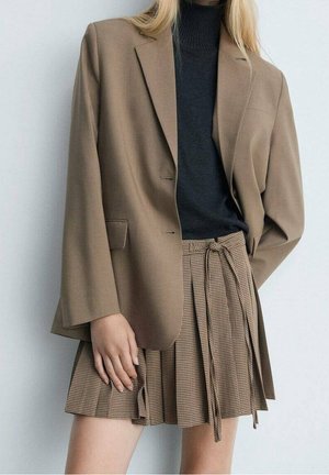Kvinde iført en brun blazer over en sort rullekrave og et plisseret, bundet, ternet brun nederdel foran en ensfarvet baggrund.