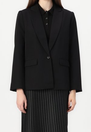 Vrouw draagt een zwarte blazer met één knoop over een zwarte overhemd met kraag en een zwarte rok met dunne witte verticale strepen.