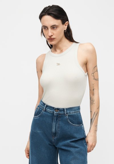 Γυναίκα με σκούρα μαλλιά που φοράει λευκό αμάνικο ριμπ τοπ και ψηλόμεσα μπλε τζιν, εμφανίζοντας τατουάζ στα χέρια της πάνω σε απλό φόντο.