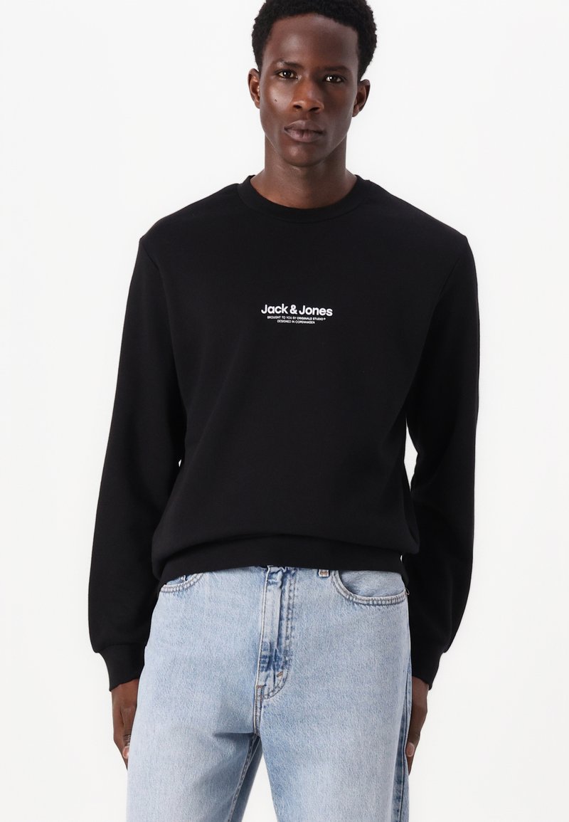 Schwarzer Sweatshirt mit Rundhalsausschnitt, mit weißem Logo-Text auf der Brust. Kombiniert mit hellblauen Denim-Jeans. Glatte Stoffoberfläche.