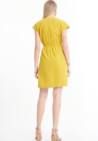 Keltainen, lyhythihainen mekko, jossa on joustava vyötärö, ulottuu polven ylle; sileä pinta; yhdistetty vaaleanbeigeihin paksupohjaisiin sandaaleihin.