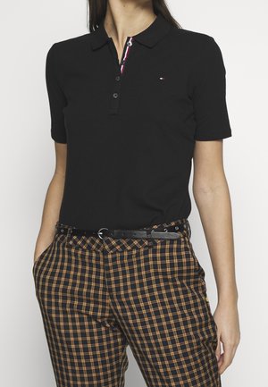 Schwarzes Poloshirt mit Kragen und kurzen Ärmeln, versehen mit einer rot-weiß gestreiften Knopfleiste. Kombiniert mit karierten schwarzen und orangefarbenen Hosen.