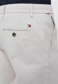 Pantaloni de culoare bej deschis cu o textură netedă, un singur buzunar posterior cu nasture și un mic logo brodat în roșu, alb și albastru.