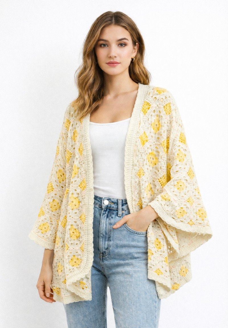 Junge Frau trägt eine cremefarbene und gelbe Häkeljacke über einem weißen Tanktop und hellblauen, hoch taillierten Jeans, sie steht vor einem weißen Hintergrund.