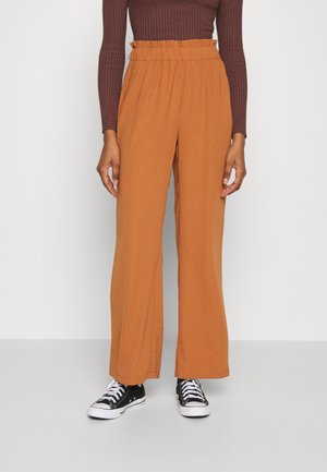 Osoba nosząca pomarańczowe, luźne spodnie z wysokim stanem, ciemnobrązową prążkowaną bluzkę z długim rękawem oraz czarne niskie trampki z białymi sznurowadłami.