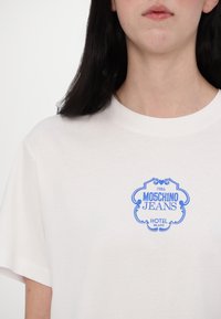 Balta kokvilnas T-krekls ar īsām piedurknēm, ar zilu logo druku, kas lasāma kā "Moschino Jeans Hotel Milano 1986" dekoratīvā fontā.