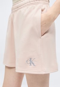 Ανοιχτό ροζ σορτς Calvin Klein με πλαϊνές τσέπες, φορεμένα με το χέρι μερικώς μέσα στην τσέπη, με το λογότυπο να είναι τυπωμένο κοντά στο τελείωμα στο αριστερό πόδι.