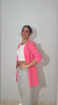 Γυναίκα που χαμογελάει, στέκεται πλαγίως φορώντας ένα φωτεινό ροζ σακάκι, λευκό κοντό τοπ και ανοιχτό γκρι τζιν ψηλόμεσο, σε απλό φόντο.