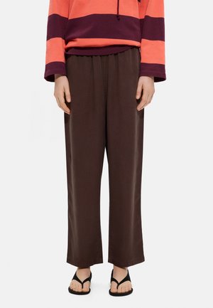 Osoba ubrana w brązowe spodnie z szerokimi nogawkami, czarne klapki i pomarańczowo-ciemnoczerwoną, paskowaną bluzkę z długim rękawem, stojąca na białym tle.