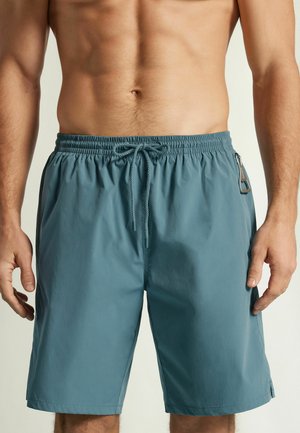 Shorts pour homme bleu sarcelle à taille élastique avec cordon de serrage et poche latérale zippée, portés par une personne au torse nu et aux bras le long du corps.