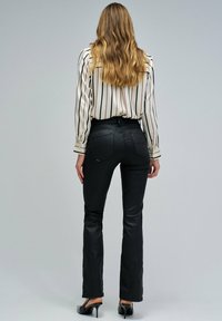Mujer con cabello ondulado y largo, vestida con pantalones ajustados negros, una blusa blanca con rayas verticales y zapatos de tacón negro, de pie de espaldas.