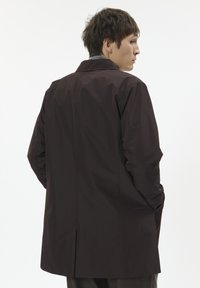 Ciemnobrązowy, wodoodporny trencz o prostym kroju, z długimi rękawami, kołnierzem z wycięciem i pojedynczym rozcięciem z tyłu. Gładka faktura.