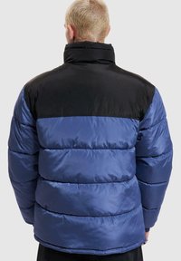 Blå och svart puffjacka med hög krage, med horisontella sömmar och en slät, glansig textur. Bakåtkik visar färgblockeringen.
