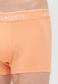 Close-up van een persoon die perzikkleurige Lacoste boxershort draagt met een gemerkte tailleband.