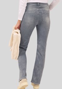 Lichtblauwe denim jeans met een rechte pijp, voorzien van twee achterzakken. Een gestructureerde crèmewitte trui wordt in de linkerhand vastgehouden.
