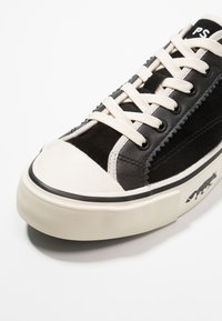 Baskets noir et blanc avec des lacets crème, un embout en caoutchouc et un logo renard sur le côté de la semelle, vues de face.