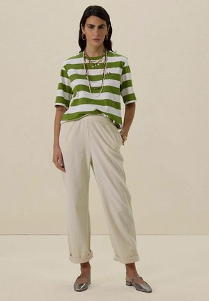 T-shirt rayé vert et blanc associé à un pantalon plissé de couleur crème. Les accessoires comprennent des colliers superposés et des chaussures argentées.