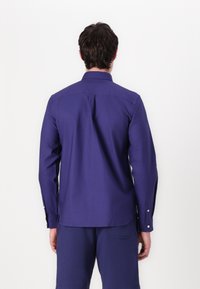 Camisa de manga larga morada, de material de algodón, con collar puntiagudo, puños abotonados, que presenta un canesú y un pliegue en la espalda para mayor movilidad.