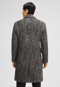 Man die met zijn rug naar de kijker staat en een knielang, gestructureerd zwart-wit visgraatjack draagt met lange mouwen en een kleine kraag.