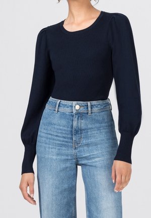 Femme portant un pull côtelé bleu marine ajusté avec des manches longues bouffantes et un jean bleu clair taille haute.