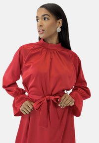 Robe rouge à manches longues en tissu lisse avec un col montant, des détails froncés et une ceinture nouée mettant la taille en valeur.