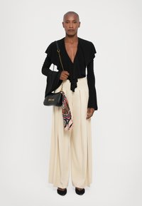 Black layered top with flared sleeves, cream wide-leg trousers, black handbag with scarf detail, and black pointed heels.