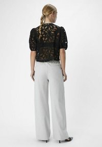 Haut en dentelle noire avec des manches bouffantes, ornée de motifs floraux, assorti à un pantalon large gris clair et des escarpins noirs pointus.