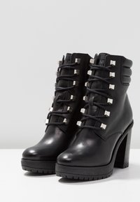 Botas de tobillo de cuero negro con suela texturizada, tacón alto grueso y acentos metálicos cuadrados en los cordones. Acabado suave y diseño resistente.