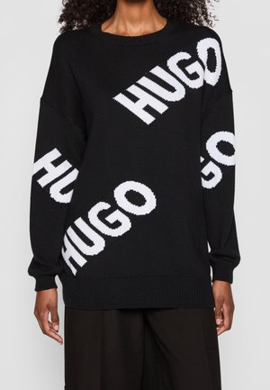 Pull tricot noir à manches longues avec un grand texte diagonal blanc "HUGO" traversant le devant et les manches, porté avec un pantalon noir par une personne.